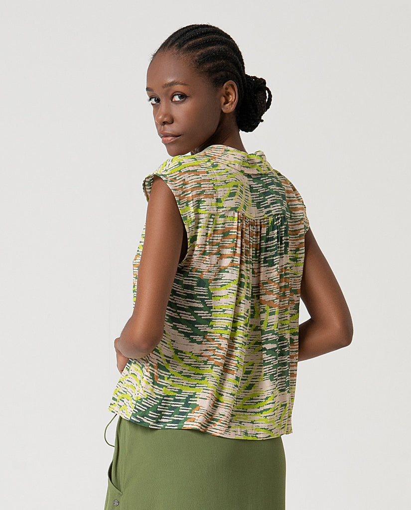 Woman wearing a sleeveless patterned top and green pants on a plain background