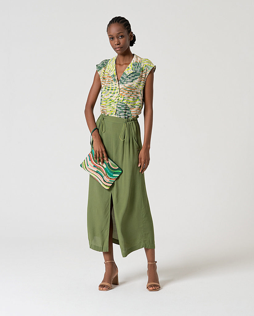 Woman wearing a patterned top and green skirt holding a matching clutch against a white background